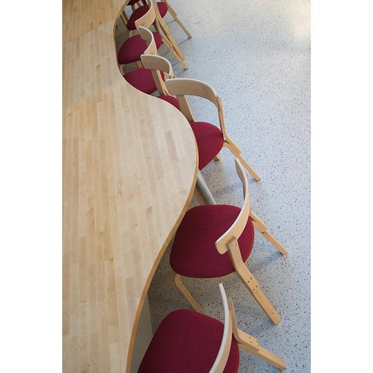 Aaltoilevan muotoinen puupintainen pöytä ja punaisilla istuintyynyillä varustetut puurunkoiset tuolit Länsikadun koulun erikoiskalusteina. Ergonomiset ja esteettiset kalusteet oppimisympäristöön.