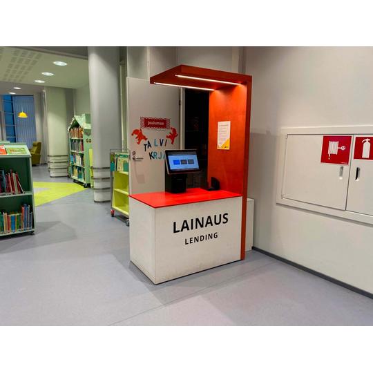 Joensuun pääkirjaston lastenosaston lainauspiste, jossa on oranssi taustaseinä ja valkoinen tiski tekstillä 'Lainaus Lending.' Tiskillä on lainausautomaatti. Taustalla näkyy kirjahyllyjä ja 'Joulumaa Talvikirjat' -koristeinen ovi.