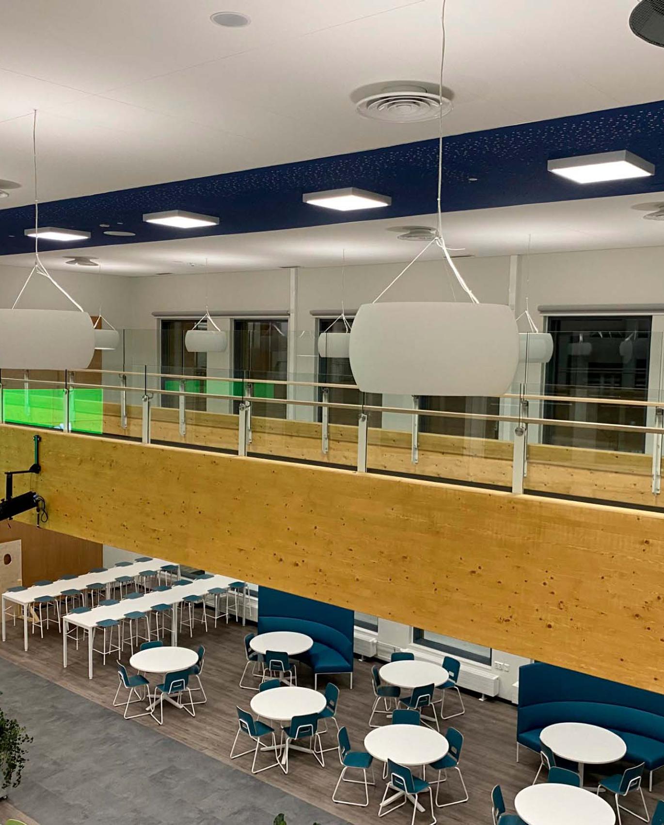 Moderni ja valoisa tilakokonaisuus, jossa on pyöreitä pöytiä, värikkäitä tuoleja ja ilmava sisustus. Ylhäällä roikkuvat valaisimet ja puinen parveke luovat avoimen ja kutsuvan ilmapiirin.