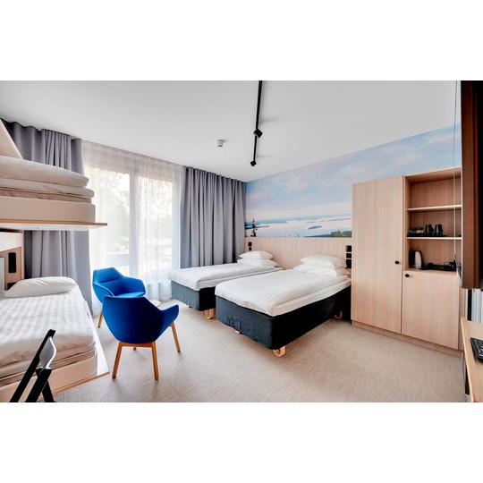 Valoisa ja moderni hotellihuone, jossa on kaksi sänkyä, kerrossänky, siniset nojatuolit ja taustaseinällä maisematapetti, joka esittää järvimaisemaa. Huoneessa on myös puinen kaapisto ja hyllytila, jossa on kahvinkeittovälineitä. Kuva: PKO.