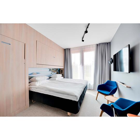 Hotellihuone, jossa kaksi erillistä sänkyä vierekkäin, vaaleapuinen sisustus ja maisemakuva sängyn päädyssä. Huoneessa on siniset nojatuolit, seinään kiinnitetty televisio ja suuret ikkunat, joista näkyy ulkoinen maisema. Kuva: PKO.