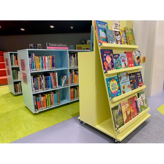 Joensuun p&auml;&auml;kirjaston lastenosaston tietokirjallisuuden osasto, jossa on sinisi&auml; hyllyj&auml; kirjoille ja vaaleankeltainen esittelyhylly suosituimmille tietokirjoille. Hyllyjen ymp&auml;rill&auml; on v&auml;rik&auml;s lattia, ja taustalla n&auml;kyy punaisia istuinportaita.