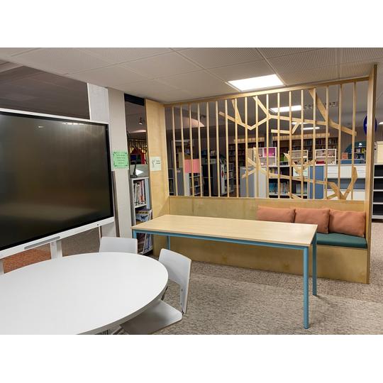 Länsikadun koulun oppimistila, jossa moderni interaktiivinen näyttö, pyöreä pöytä valkoisilla tuoleilla, sekä penkki ja pöytä, jotka on erotettu puukuviollisella väliseinällä. Taustalla näkyy hyllyjä täynnä kirjoja.