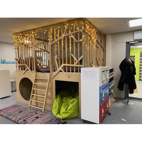 Länsikadun koulun leikkialue, jossa on puinen mökki oksakuvioisilla kaiteilla, koristelamppuja, tikkaat, pehmeitä istuintyynyjä ja värikäs matto.