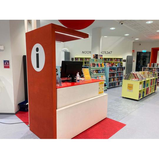 Joensuun p&auml;&auml;kirjaston lastenosaston infopiste, jossa on punainen taustasein&auml; ja p&ouml;yt&auml;, jossa on tietokone, k&auml;sidesi ja opastetauluja. Pisteen edess&auml; on punainen lattiaosio, ja taustalla n&auml;kyy hyllyj&auml; t&auml;ynn&auml; nuortenkirjoja.