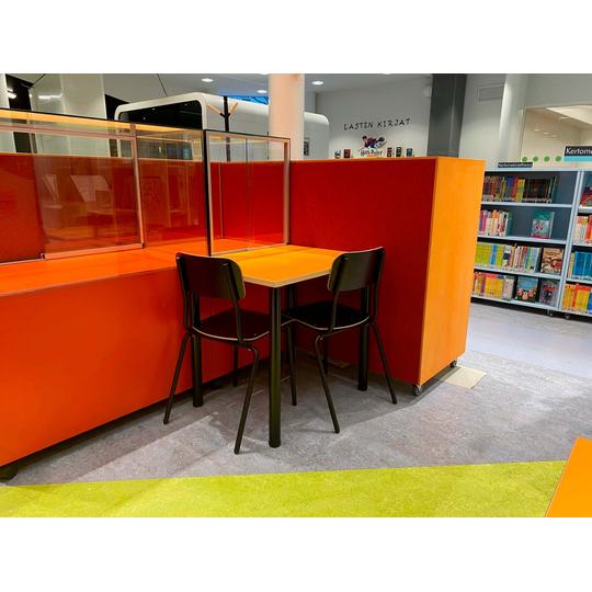 Joensuun pääkirjaston lastenosaston oranssinsävyinen työskentelypiste, jossa on pöytä ja kaksi tuolia. Taustalla näkyy lasten kirjallisuutta sisältäviä hyllyjä ja seinällä 'Lasten kirjat' -teksti.