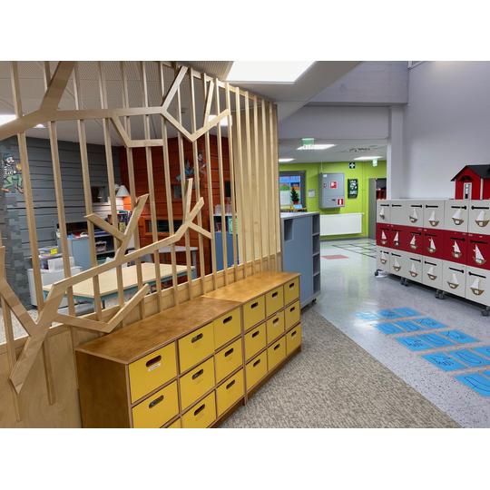 Länsikadun koulun värikäs oppimisympäristö, jossa näkyy puisia säilytyslaatikoita, leikittelevä väliseinä oksamaisilla muodoilla, sekä punaisia ja valkoisia säilytyskaappeja purjeveneaiheisilla kuvioilla.