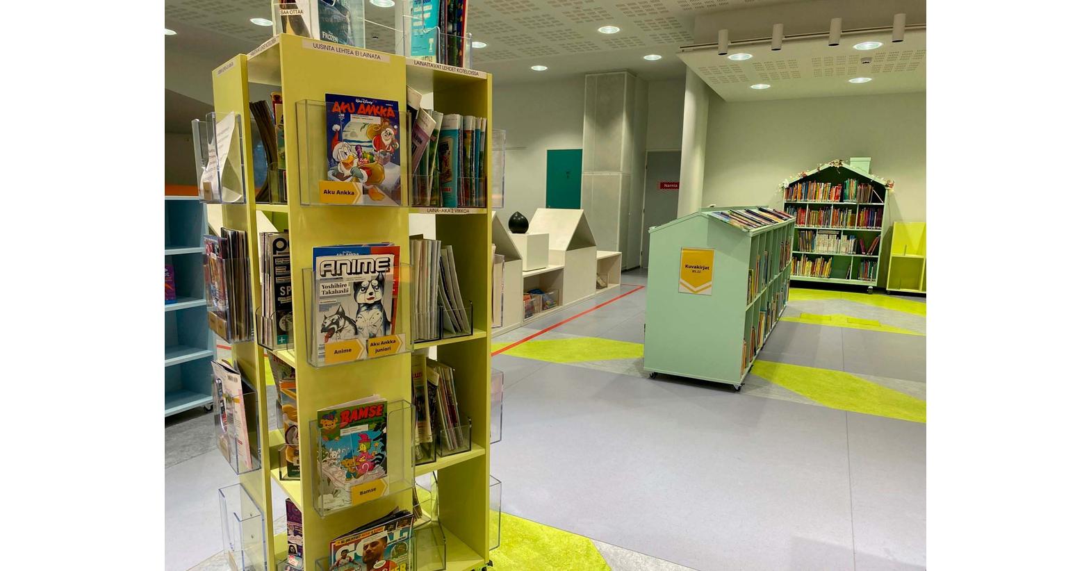 Joensuun pääkirjaston lastenosasto, jossa on värikkäitä hyllyjä täynnä kirjoja ja lehtiä. Etualalla keltainen lehtihylly ja taustalla näkyy kirjahyllyjä sekä värikkäitä kuvioita lattiassa.