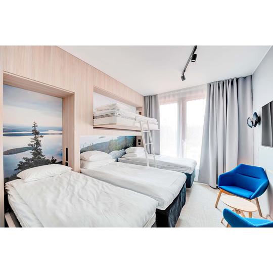 Hotellihuone, jossa kolme yksittäistä sänkyä ja yksi yläsänky tikkailla. Sängyn päädyissä on maisemakuvat, ja huoneen sisustus on vaaleapuinen. Huoneessa on sininen nojatuoli ja suuret ikkunat, joista avautuu valoisa näkymä. Kuva: PKO.