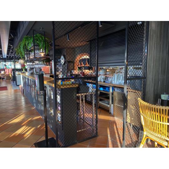 Rosso Joensuun tyylik&auml;s baaritiski, jossa yhdistyv&auml;t puupinta ja metalliverkkoelementit, sek&auml; katosta riippuvat viinilasitelineet ja vehre&auml; viherkasvi.