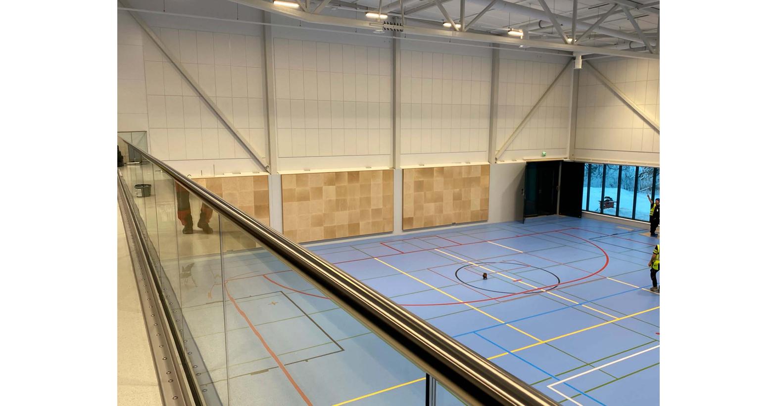 Lehmon koulun liikuntasali Kontiolahdella, jossa on vaaleansiniset lattiamerkinnoillä varustetut urheilulattiat, seinälle asennetut akustiset paneelit ja suuret ikkunat, jotka tuovat luonnonvaloa sisään. Kuvassa näkyy myös lasikaiteen yläosa katsomotasolta.