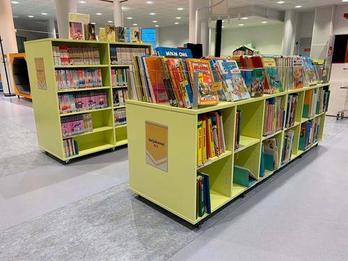 Kirjaston lastenosasto, jossa on vaaleankeltaisia kirjahyllyj&auml; t&auml;ynn&auml; v&auml;rikk&auml;it&auml; sarjakuvia ja lastenkirjoja, modernissa ja valoisassa ymp&auml;rist&ouml;ss&auml;.