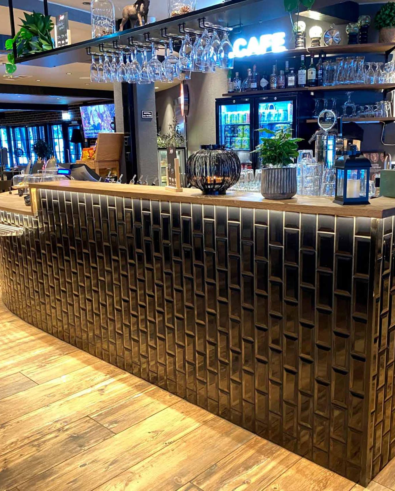 Tyylik&auml;s ja moderni baari, jossa mustat laatat, puinen tiski ja hyllyt t&auml;ynn&auml; laseja sek&auml; kasveja. Taustalla n&auml;kyy baarin ymp&auml;rist&ouml; ja sisustus.