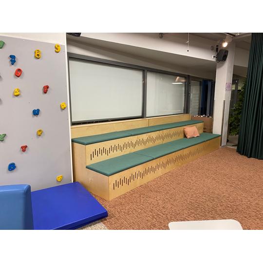 Länsikadun koulun monitoimitila, jossa on kiipeilyseinä, puiset istuinportaat vihreillä pehmusteilla ja yksittäisellä tyynyllä sekä vaaleanruskea kokolattiamatto.
