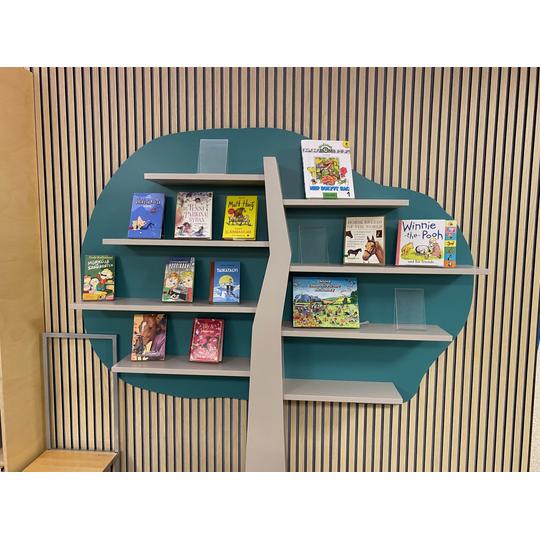 Seinälle kiinnitetty moderni vihreä hyllyjärjestelmä, jossa on erilaisia lasten- ja nuortenkirjoja. Hyllyn taustalla pystylaudoitus lisää tilan visuaalista ilmettä.