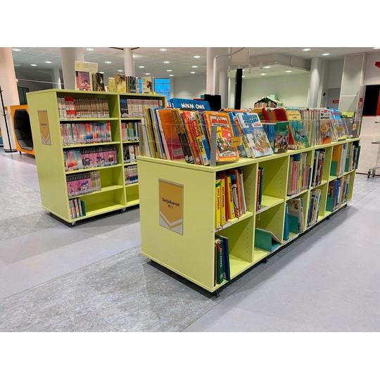 Joensuun p&auml;&auml;kirjaston lastenosaston sarjakuvahyllyt, joissa on vaaleankeltaisia siirrett&auml;vi&auml; hyllyj&auml; t&auml;ynn&auml; sarjakuvia, kuten Aku Ankka ja muita suosittuja lastenlehti&auml;. Taustalla n&auml;kyy kirjaston muita osastoja ja v&auml;rik&auml;st&auml; sisustusta.