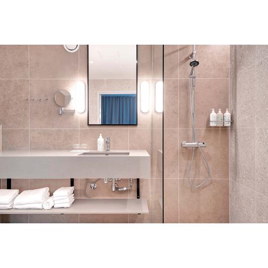 Moderni kylpyhuone, jossa beige-laatoitus, minimalistinen allas ja suuri peili. Peilin molemmin puolin on valaisimet, ja suihkussa on krominen sadesuihku sek&auml; hyllyll&auml; shampoo- ja saippuapulloja. Alahyllyll&auml; valkoisia pyyhkeit&auml;. Kuva: PKO.