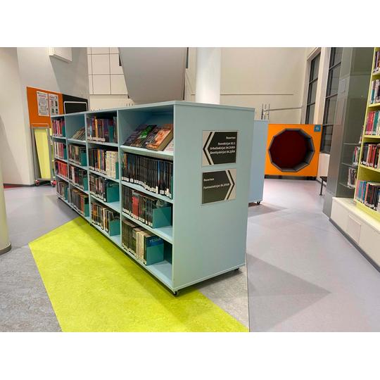 Joensuun p&auml;&auml;kirjaston lastenosaston vaaleansininen hylly, jossa on kirjoja nuorille, kuten runokirjoja, urheilukirjoja, j&auml;nnityskirjoja ja fantasiakirjoja. Taustalla n&auml;kyy kirjaston v&auml;rik&auml;s sisustus, mukaan lukien oranssi lukunurkkaus kahdeksankulmion muotoisella aukolla.