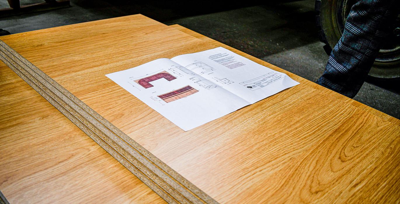 Puupintaisten materiaalien ja suunnitelmien tarkastelu ty&ouml;pajassa. Kuvassa n&auml;kyy puupinta ja sen p&auml;&auml;ll&auml; piirustukset, jotka liittyv&auml;t suunnitelmaan.