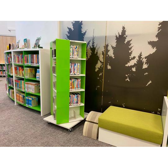 Karsikon kirjaston lasten osasto, jossa on vihre&auml;-sohva, v&auml;rikk&auml;it&auml; kirjoja sis&auml;lt&auml;vi&auml; kirjahyllyj&auml; ja py&ouml;rill&auml; liikkuva vihre&auml; hylly. Taustalla sein&auml;ss&auml; kuusimets&auml;n siluettikoriste.