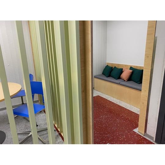 Vaaleanvihreillä väliseinillä rajattu pieni oppimistila, jossa on pyöreä pöytä sinisine tuoleineen ja viihtyisä istuinalue, joka on varustettu värikkäillä tyynyillä. Punainen lattia tuo tilaan lämpöä ja kontrastia.