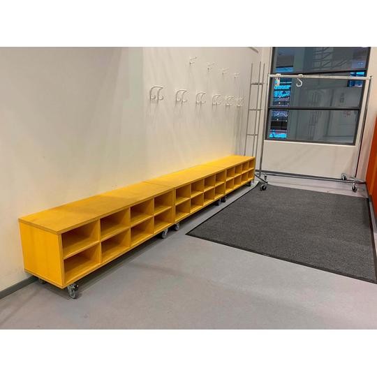 Joensuun p&auml;&auml;kirjaston lastenosaston keltainen s&auml;ilytyskaluste, jossa on avohyllyt, sek&auml; viereinen metallinen vaateteline py&ouml;rill&auml;. Taustalla sein&auml;&auml;n kiinnitettyj&auml; koukkuja ja ikkuna.