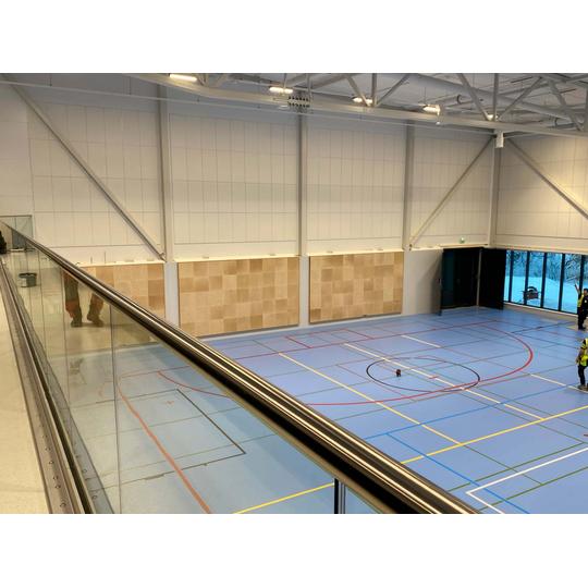 Lehmon koulun liikuntasali Kontiolahdella, jossa on vaaleansiniset lattiamerkinnoill&auml; varustetut urheilulattiat, sein&auml;lle asennetut akustiset paneelit ja suuret ikkunat, jotka tuovat luonnonvaloa sis&auml;&auml;n. Kuvassa n&auml;kyy my&ouml;s lasikaiteen yl&auml;osa katsomotasolta.