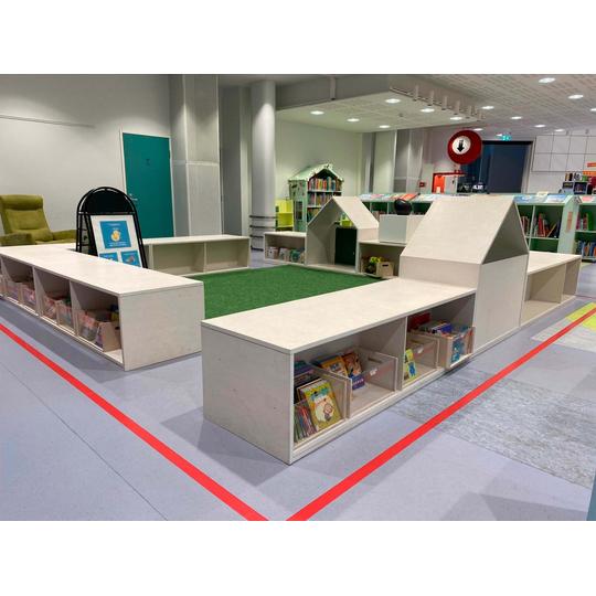 Joensuun pääkirjaston lastenosaston matalat, valkoiset hyllyelementit, jotka muodostavat talon muotoisia rakenteita. Keskellä on vihreällä tekonurmella päällystetty alue, ja hyllyissä on sijoitettuna selkeästi merkityt laatikot, joissa on erilaisia lastenkirjoja.