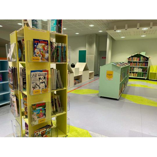 Joensuun p&auml;&auml;kirjaston lastenosasto, jossa on v&auml;rikk&auml;it&auml; hyllyj&auml; t&auml;ynn&auml; kirjoja ja lehti&auml;. Etualalla keltainen lehtihylly ja taustalla n&auml;kyy kirjahyllyj&auml; sek&auml; v&auml;rikk&auml;it&auml; kuvioita lattiassa.