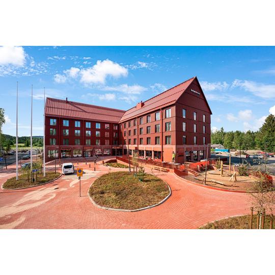 Break Sokos Hotel Koli Kyl&auml;n punainen rakennus, jonka edess&auml; on laatoitettu piha-alue ja viheristutuksia. Piha-alueella on leikkipaikka, istutuksia ja pys&auml;k&ouml;ityj&auml; autoja, taustalla n&auml;kyy mets&auml;maisema. Kuva: PKO.