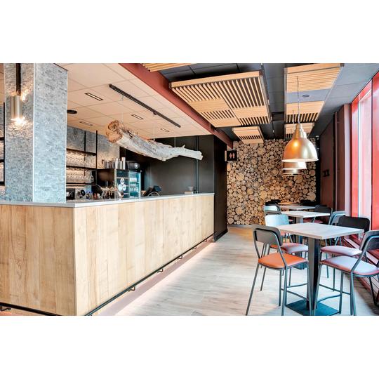 Ravintolan tyylikäs sisustus, jossa yhdistyvät luonnonmateriaalit ja moderni muotoilu. Puiset kalusteet ja katosta roikkuva puunrunko luovat lämpimän tunnelman. Taustaseinä on koristeltu halkopinolla, ja valaistus tuo tilaan kodikkuutta. Kuva: PKO.