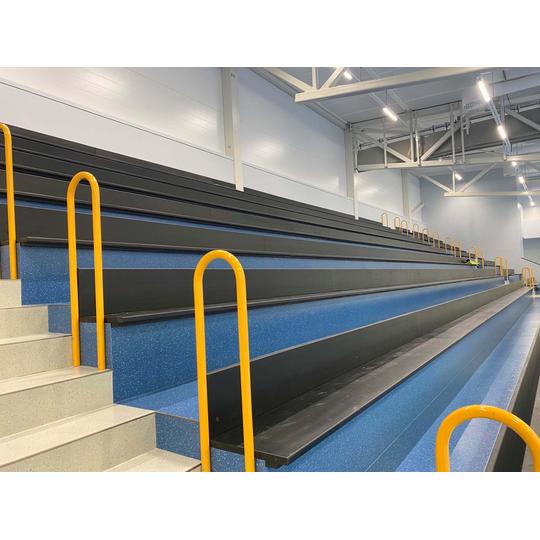 Lehmon koulun liikuntasalin katsomo Kontiolahdella, jossa on siniset portaikot, mustat istuinpenkit ja keltaiset kaiteet. Katsomon taustalla n&auml;kyy vaaleat sein&auml;t ja kattorakenteet, jotka korostavat avaraa tilaa.